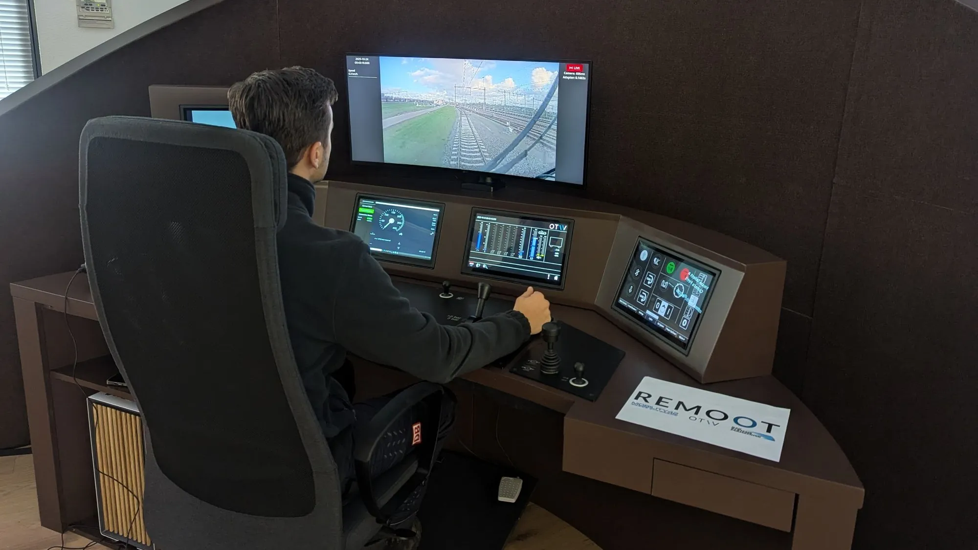 An operator at OTIV's REMOOT desk driving a train remotely: a live forward track view on the upper screen, with speed, brake and control instruments on the screens below.